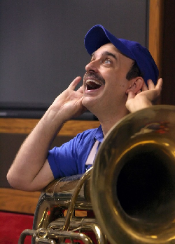Mr. Kippy with a tuba in his lap, holding his ears